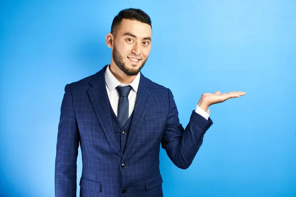 Asian Kazakh man in a business suit with a tie happily pretends to be holding something on his palm against a blue background