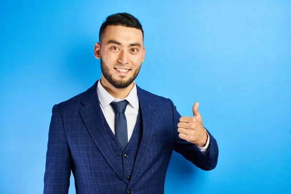 Successful Asian Kazakh businessman shows thumbs up and toothy smiles on a blue studio background. Isolated