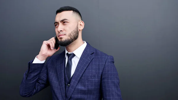 Asian Kazakh businessman speaks on a mobile phone with a serious expression on the black wall background.