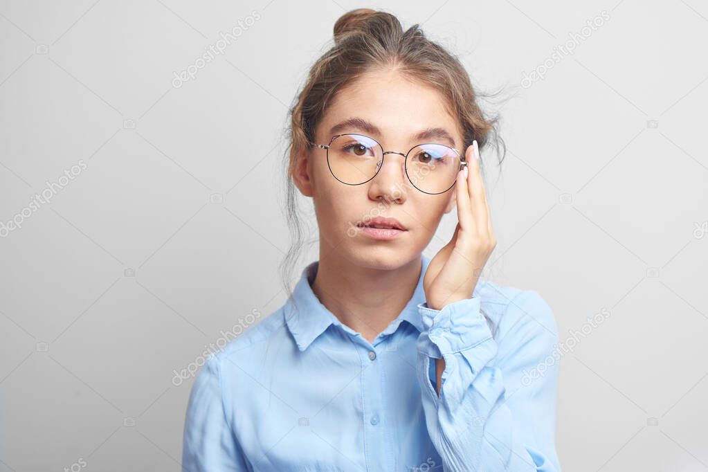 Portrait of pensive Asian Kazakh young woman with bun of hair on head in stylish glasses wearing a shirt in white studio, thinking girl