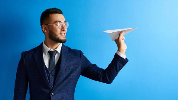 Smiling Asian Kazakh businessman in suit and glasses holds paper airplane in hand on blue studio background. Small business development concept, project launch, team training, start-ups, global strategy