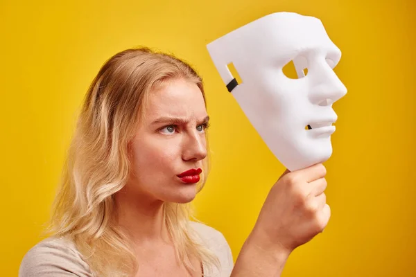 Emotional female person with white mask on yellow background. Internet fraud concept, anonymous, incognito, bipolar personality disorder, hypocris
