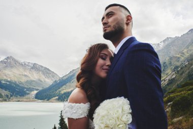 Dağ gölünde güzel düğün fotoğrafı. Aşık Mutlu Asyalı çift, beyaz elbiseli gelin ve takım elbiseli damat Kazak manzara arka plan karşı fotoğraflandı