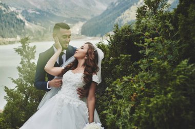 Dağ gölünde güzel düğün fotoğrafı. Aşık Mutlu Asyalı çift, beyaz elbiseli gelin ve takım elbiseli damat Kazak manzara arka plan karşı fotoğraflandı