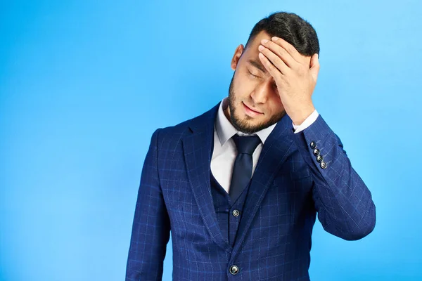 Asian Kazakh man in business suit holds his head, experiencing stress and headache at office work, tired of fuss isolated on blue studio background