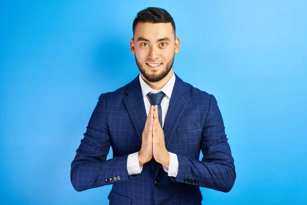 Friendly Asian Kazakh man in business suit smiling clasped his hands in thankful prayer pose isolated on blue background