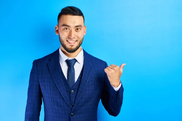 Asian Kazakh man in a business suit smiles friendly and shows finger to the sides on copy space on a blue wall isolated