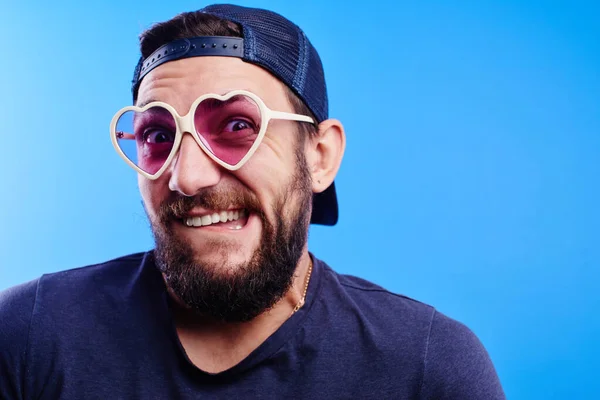 Cool funny Caucasian bearded guy in cap and glasses with hearts playfully bit his lip, with facial expression like oops! isolated on blue background