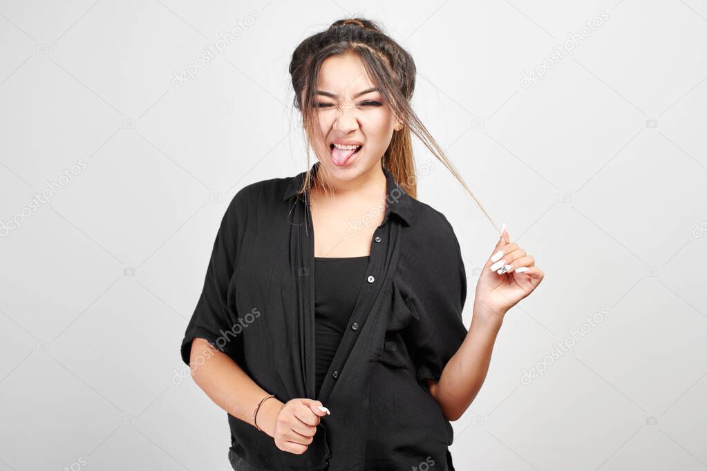 Asian Kazakh girl in total black on a white background playfully shows the tongue and touch hair, studio portrait