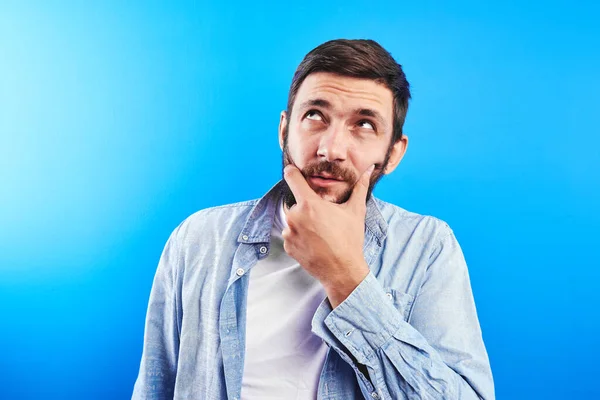 Puzzled caucasian bearded man with pensive face looks up holds chin isolated on blue studio background