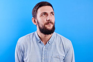 Portrait of fantasizing handsome caucasian bearded man in casual with serious face looks up dreams and imagines isolated in blue studio