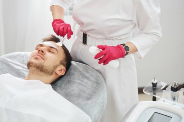 Güzellik salonundaki kozmetoloji prosedürlerinde çalışan yakışıklı beyaz bir genç adam. Erkek dermatolog cilt bakımı, yüz maskesi.