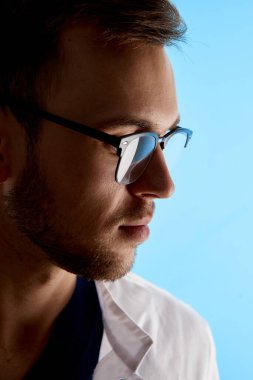 Mavi duvarda izole edilmiş ciddi bir yüz ifadesi olan, beyaz önlüklü ve gözlüklü doktorun profil portresi.
