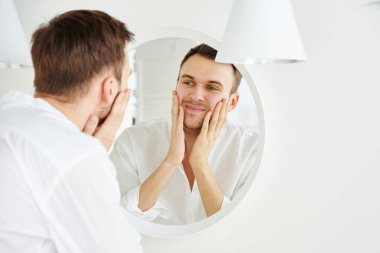 Genç, çekici, beyaz bir adam dokunuyor, aynada yüzünün tadını çıkarıyor ve yakından gülümsüyor. Cilt bakımı kavramı