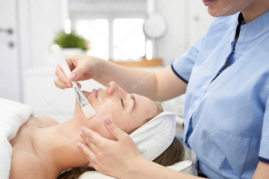Doctor cosmetologist makes a multi-stage facial cleansing procedure to a young attractive woman in a beauty salon. Summer skin care