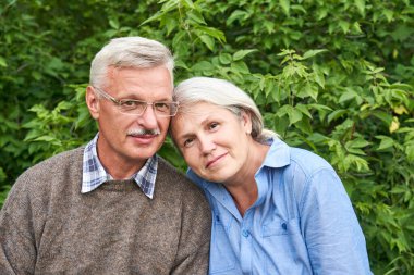 Birbirine aşık yaşlı bir çift parkta bir bankta oturuyor, gülümsüyor ve yeşil ağaçların arkasında sarılıyorlar.