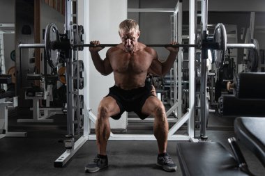 Kaslı, yetişkin, vahşi bir adam spor salonunda halterli çömelme yapıyor. Kafkas özgün vücut geliştirme egzersizlerinin portresi.