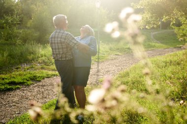 Bir yaz parkında romantik bir randevuya çıkmış yaşlı bir çift yeşil ağaçların arkasında gülümsüyor ve sarılıyor.