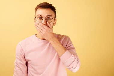 Şok olmuş bir adamın portresi ağzını eliyle kapatıyor, duygusal beyaz yüzüyle pembe günlük kıyafetlerle sarı arka planda izole edilmiş.