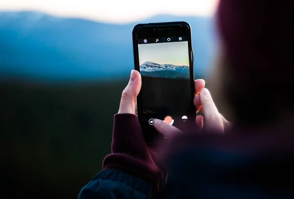 Girl taking photos of the snow-capped mountains using the phone. Close-up shot of the hand holding the phone with the mountain behind.