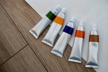 Beş canlı boya tüpü sağlam ahşap bir masanın üzerinde duruyor, çeşitli sanatsal projelerde yaratıcı kullanıma hazır ve hobi olarak yeşil turuncu mavi kahverengi renkler çiziyor.