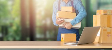 İş yerindeki dizüstü bilgisayar, küçük işletme sahibi. Müşteriye teslim etmek için karton kutu dolusu ürün. Çevrimiçi satış, e-ticaret, paketleme konsepti, sabah ışığı, evden çalışma, bulanık görüntü.
