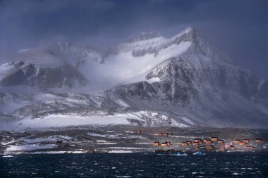 A stormy mountainous scene at the tip of the Antarctic peninsular. An Antarctic research base is seen at the foot of the mountain clipart
