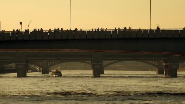 テムズ クリッパー リバーバスは 日没時にロンドン橋の下を通過します 多くの人がシルエットでロンドン橋を渡っている — ストック動画