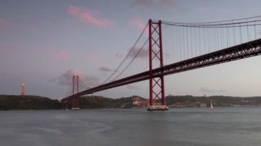 Akşam alacakaranlığı sırasında 25 Nisan Tagus Nehri Köprüsü 'nün alçak ve geniş açılı klibi. Arka planda İsa heykeli var ve altında küçük bir yat geçiyor.