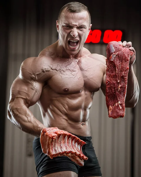 Big muscle handsome pretty young blonde man violent butcher athlete bodybuilder chopping pork and beef meat with axe screaming with anger and frowning. Predator concept. Fitness man at workout in gym