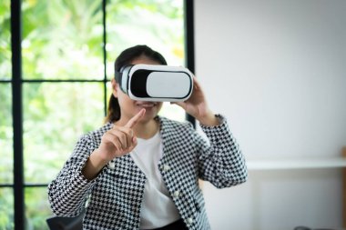 Siyah beyaz ceket giyen bir kadın sanal gerçeklik kulaklığıyla bir video oyunu oynuyor. Ekrandaki bir şeyi işaret ediyor.