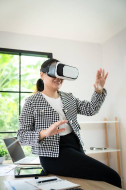 Siyah beyaz kareli ceket giyen bir kadın önünde bir tabletle bir masada oturuyor. O, beyaz bir gömlek ve siyah pantolon giyiyor. Sanal gerçeklik kulaklığı takıyor.