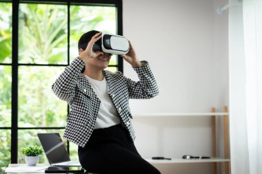 Siyah beyaz ceket giyen bir kadın dizüstü bilgisayarın önünde oturuyor ve elinde sanal gerçeklik kulaklığı var. Gülümsüyor ve sanal gerçeklik deneyiminin tadını çıkarıyor.