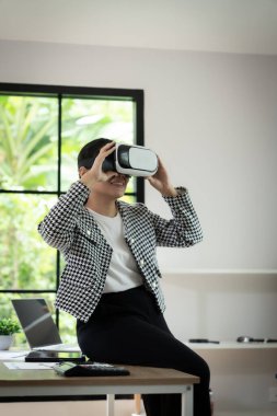 Siyah beyaz ceket giyen bir kadın bir masada dizüstü bilgisayar ve tabletle oturuyor. VR kulaklığı takıyor ve bu deneyimden zevk alıyor.