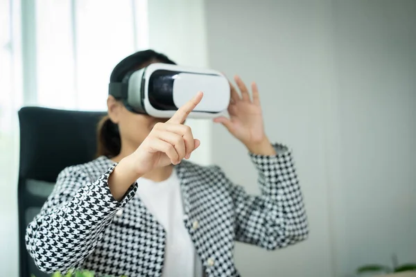 Siyah beyaz ceket giyen bir kadın sanal gerçeklik oyunundaki bir şeyi işaret ediyor. Sahne, bir sandalye ve saksı bitkisi olan bir odada geçiyor.