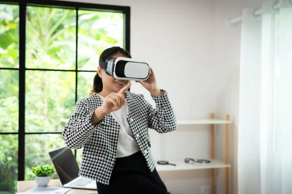 Siyah beyaz ceket giyen bir kadın sanal gerçeklik kulaklığıyla bir video oyunu oynuyor. Ekrandaki bir şeyi işaret ediyor ve oda saksı bitkisi ve dizüstü bilgisayarla süslenmiş.