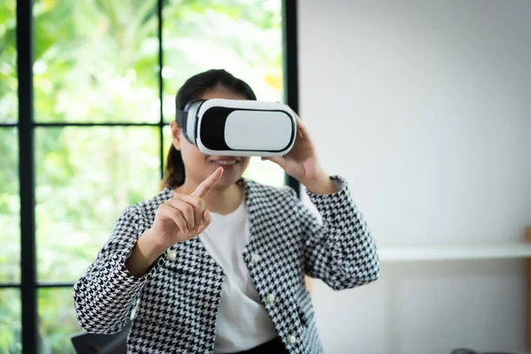 Siyah beyaz ceket giyen bir kadın sanal gerçeklik kulaklığıyla bir video oyunu oynuyor. Ekrandaki bir şeyi işaret ediyor.