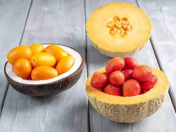 Vista de cerca de la fresa en melón y kumquat en un tazón de coco y media de melón sobre fondo de madera blanca — Foto de Stock