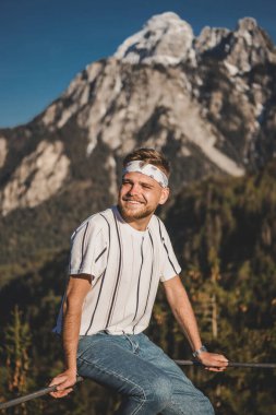 Genç hippi adam tırabzanda oturur ve kaya dağı ormanının arka planında gülümser. Dikey portre