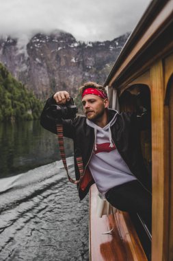 Hipster man shoots on camera from the window of the wooden boat on background of lake Konigssee & rock mountain in clouds. Bavaria. Germany clipart