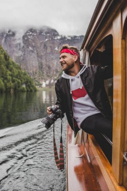 Konigssee 'de. Bavyera 'da. Almanya. Mutlu hippi çocuk ahşap teknenin penceresinde oturur ve gölün arka planında kameralar ve gülücükler, bulutlarda orman ve kaya dağları vardır.