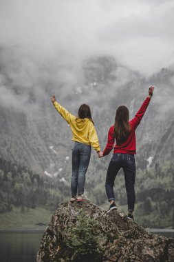 Sarı ve kırmızı tişörtlü iki kız Obersee Gölü, Rothbach Şelalesi, bulutlu havada kaya dağı ve yeşil ormanın arka planındaki taşta özgürlüğü hissediyorlar. Bavyera 'da. Almanya. Dikey duvar kağıdı