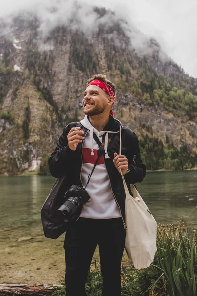 Hipster adam kamera ve çanta ile duruyor ve Konigssee Gölü 'nün arka planında sigara tüttürüyor ve bulutlar içinde kaya dağı. Bavyera 'da. Almanya