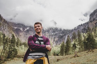 Obersee 'de. Bavyera 'da. Almanya. Genç esmer adam ormanın arka planında şelaleler, dağlar, çamlar ve bulutlarda gülümsüyor.