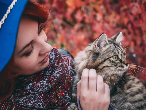 Beautiful red-haired woman in blue hat and leather jacket walking with cat in autumn red park. — Stock Photo, Image