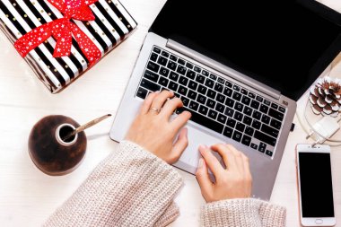 İş yerinde dizüstü bilgisayarda çalışan bir kadın. Kadın eli, dizüstü bilgisayar, akıllı telefon, şarj aleti, paketlenmiş hediye, arkadaşla su kabağı, ofis veya serbest kış tatili konsepti