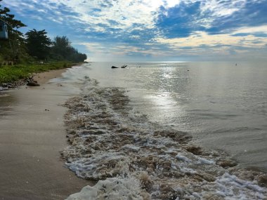Küçük kahverengi köpük dalgaları bulutlu bir gökyüzünün altında kumlu bir plaja vuruyor..