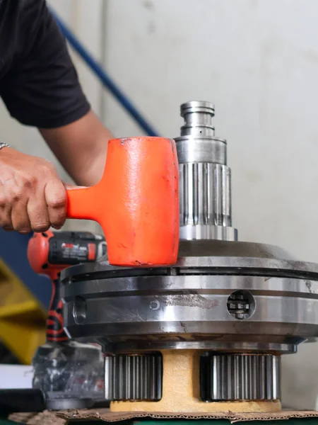 İşçi, atölyede hassas dişleri olan büyük metal teçhizat montajını ayarlamak için turuncu lastik çekiç veya yumuşak çekiç kullanıyor..