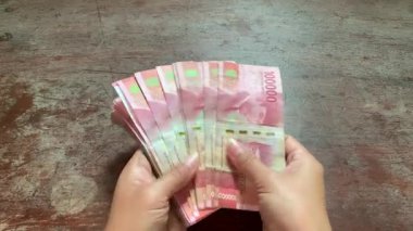 Hands spread and checking Indonesian rupiah banknotes under natural light on rustic table. Detailed 4K shot, perfect for finance, savings, fraud, security, and business themes.