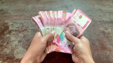 Hands spread and checking Indonesian rupiah banknotes under natural light on rustic table. Detailed 4K shot, perfect for finance, savings, fraud, security, and business themes.